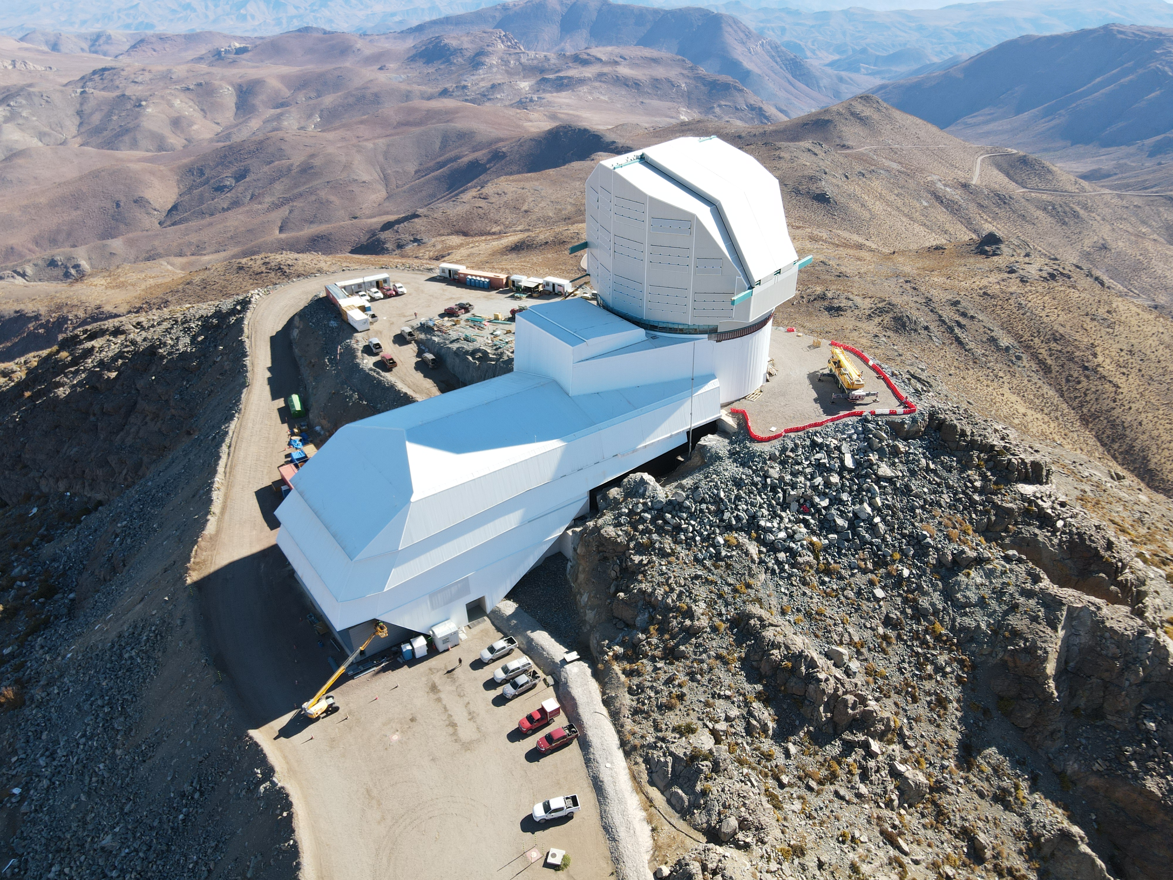 Photo of Rubin Observatory taken from above by a drone. The observatory has a white dome and a long support building that extends to the left of the dome. The observatory is on a dry, brown mountain top with other mountains stretching into the distance.